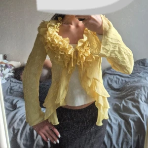 Gul volangig blus med knyt - Supersöt gul blus med volanger längs hela urringningen och framtill. Lätt transparent material, långa ärmar och knyt framtill. Perfekt för dig som vill sticka ut med en romantisk och lekfull look. Sitter figurnära över midjan och har flowiga ärmar. Den har 3 st knappar, samt ett knytband. Står ingen storlek på men jag har xs-s så den passar någon med den storleken men kan nog även passa M då den inte är tajt av sig. 