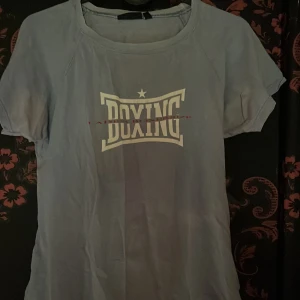 Ljusblå BOXING t-shirt från Melly - Ljusblå t-shirt från Melly med trycket 'BOXING' i vitt framtill. Klassisk rund halsringning och korta ärmar. Perfekt för dig som gillar sportiga vibes och enkel stil.