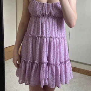 Ljuslila blommig volangklänning - Supersöt ljuslila kortklänning med små rosa blommor, volanger i flera lager och smock i ryggen. Axelbanden är tunna och knyts upptill. Perfekt flowy modell för varma dagar. Klänningen är ifrån newyorker och har defekt på sidan av bröstet (se bild). Jag själv märker knappt defekten❤️