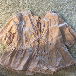 Ljusrosa blus med knyt och volang - Supersöt ljusrosa blus med korta ärmar, volang nertill och två knyt framtill. Blusen har en luftig passform och lätt glansigt tyg som ger en romantisk vibe. Perfekt att styla med jeans eller kjol för en trendig look.