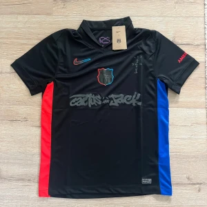 FC Barcelona x Cactus Jack Nike tröja - Snygg svart FC Barcelona x Cactus Jack fotbollströja från Nike. Tröjan har korta ärmar, röda och blå sidopaneler, klubbmärke på bröstet och coolt Cactus Jack-tryck framtill. Materialet är lätt och andas, perfekt för fotboll eller streetwear.