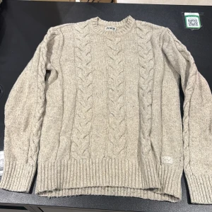 Beige kabelstickad tröja från Edwin - Kabelstickad beige tröja från Edwin med grov struktur och rund hals. Tröjan har ribbade muddar vid ärmslut och nederkant samt ett diskret Edwin-märke nertill. Materialmix med ull och bomull gör den både mjuk och varm. Perfekt för lager-på-lager under kyliga dagar.