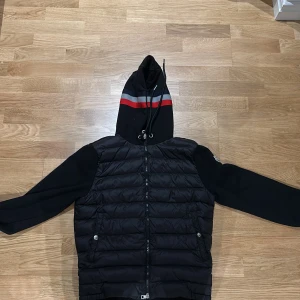 Svart dunväst med huva från Moncler - Svart dunväst från Moncler med quiltad front, hel dragkedja och två fickor med tryckknappar. Västen har en svart huva med grå och röd rand samt dragsko. Ärmarna är i mjuk bomull och vänster ärm har Moncler-logga. Perfekt för lager-på-lager-stil.