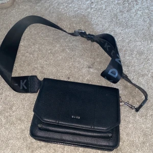 Svart axelväska från NA-KD - Snygg svart axelväska från NA-KD i stilren design med bred justerbar axelrem med logga. Väskan är tillverkad i strukturerat skinnimitation och har ett lock med diskret silverfärgad logotyp framtill. Perfekt storlek för det viktigaste och passar till många outfits. Har lite slit på fram sidan se bilderna! 