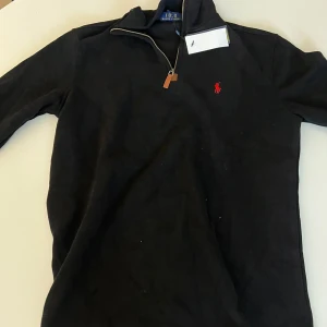Svart half zip tröja från Ralph Lauren - Svart långärmad tröja från Polo Ralph Lauren med half zip och klassisk röd logga broderad på bröstet. Tröjan har hög krage och är tillverkad i mjuk bomull, perfekt för en clean och stilren look.