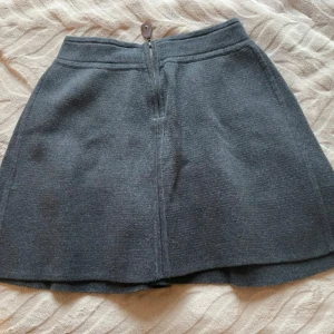 Mörkgrå stickad kjol från Newhouse - Mörkgrå stickad kortkjol från Newhouse med snygg A-linjeform och markerad midja. Kjolen har dragkedja bak med en brun läderdetalj och dekorativ knapp. Perfekt för dig som gillar klassisk och enkel stil. 100% bomull