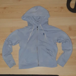 Ljusblå hoodie från Polo Ralph Lauren - Ljusblå hoodie från Polo Ralph Lauren med dragkedja, huva och vita snören. Klassisk broderad logga på bröstet och känguruficka framtill. Tillverkad i mjuk bomull, perfekt för chill dagar eller när du vill ha en clean look. Några små skador. Mott från axeln till midjan 50 centimeter 