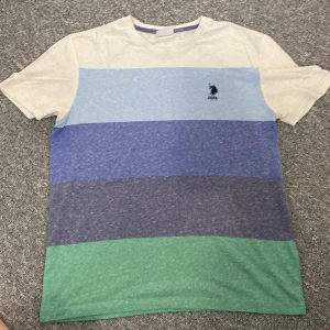 Randig t-shirt från U.S. Polo Assn. - U.S Polo Assn. med randig mönster. Snygg T-shirt men har dock ett litet hål vid botten som man kan se i sista bilden. Vid frågor eller funderingar hör av dig!