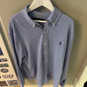 Blå skjorta från Ralph Lauren - Snygg blåvit rutig skjorta från Ralph Lauren med klassisk button-down krage och broderad logga på bröstet. Skjortan har långa ärmar och knäppning framtill. Perfekt för dig som gillar stilrena och tidlösa plagg.