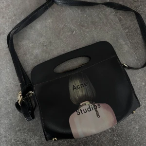 Svart axelväska Acne Studios - Cool svart axelväska från Acne Studios i skinn med unik print av en person bakifrån och texten Acne Studios. Väskan har både handtag och justerbar axelrem, guldfärgade metalldetaljer och dragkedja upptill. Perfekt för dig som gillar statement-accessoarer.