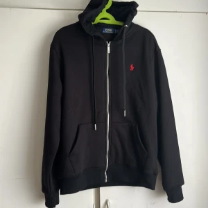 Svart hoodie från Polo Ralph Lauren - Svart hoodie från Polo Ralph Lauren med dragkedja, huva och snörning. Klassisk röd broderad logga på bröstet och två fickor framtill. Tillverkad i mjuk bomull, perfekt för chill dagar. Snygg och enkel att matcha med allt.