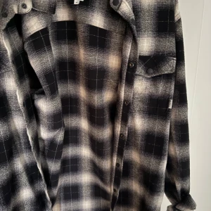 Svartvit rutig overshirt - Snygg overshirt i svart och vitt med rutigt mönster och knappar framtill. Skjortan har bröstficka med lock och klassisk krage. Perfekt att slänga över en t-shirt för en avslappnad look.