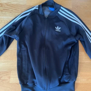 Svart adidaströja - Svart Adidaströja med klassiska vita ränder längs ärmarna och broderad logga på bröstet. Tröjan har dragkedja framtill, två fickor och ribbade muddar. Perfekt för en sportig och avslappnad stil.