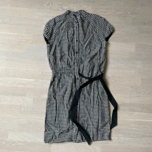 Rutig kortklänning från Moschino - Supersöt svartvit rutig kortklänning från Moschino Cheap and Chic. Klänningen har korta ärmar, knappar framtill, markerad midja med svart knytband och klassisk krage. Perfekt för dig som gillar en lekfull och trendig stil.