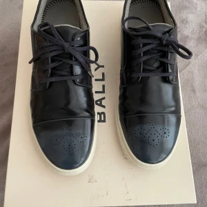 Svarta sneakers från Paul Smith - Stilrena svarta sneakers från Paul Smith i skinn med vita sulor. Skorna har klassisk snörning, rund tå och dekorativa perforeringar på tån. Insidan är ljusgrå med diskret mönster. Perfekta för dig som gillar en clean och modern look.