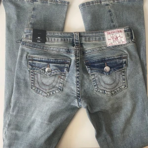 True Religion Joey Low Rise Flare Jeans - Snygga ljusblå jeans från True Religion, modell Joey Low Rise Flare. De har låg midja, utsvängda ben och klassiska kontrastsömmar. Bakfickorna har lock och silverfärgade knappar med logga. Jeansen är i denim med slitningar för en cool look. Byxorna är helt nya och har endast provats på. 
