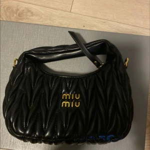 Svart quiltad axelväska från Miu Miu - Säljer en svart quiltad axelväska från Miu Miu i skinn med guldiga detaljer och logga framtill. Väskan har ett mjukt, puffigt mönster och dragkedja upptill. Perfekt storlek för det viktigaste och en snygg accessoar till din outfit.