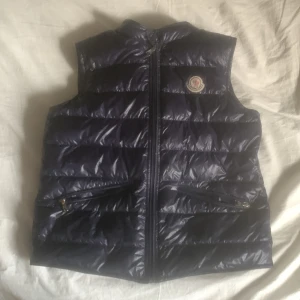 Mörkblå dunväst från Moncler - Säljer en mörkblå dunväst från Moncler med klassisk quiltad design och ikonisk logotyp på bröstet. Västen har glansig yta och är perfekt för lager-på-lager. Fodrad med dun för extra värme och stilren look.