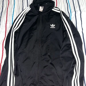 Svart Adidas originals kofta  - Klassisk svart Adidas originals kofta  med dragkedja och de ikoniska tre vita ränderna längs ärmarna. Jackan har Adidas logga på bröstet och är tillverkad i polyester. Perfekt för en sportig och avslappnad stil.