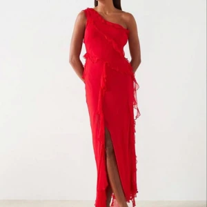 Röd långklänning med volang och slits - Snygg röd långklänning i luftig polyester med volangdetaljer och en axel. Klänningen har en slits framtill och asymmetrisk oneshoulder-skärning som ger en cool och trendig look. Perfekt för dig som vill sticka ut med en färgstark och elegant stil. Nypris ca 900kr