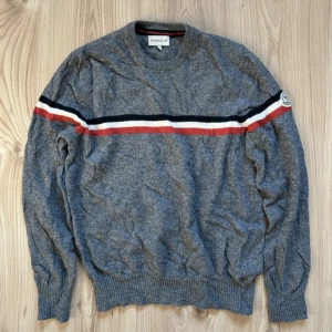 Grå Moncler stickad tröja  - Snygg grå stickad tröja från Moncler i storlek L i barnstorlek, passar som XS! Tröjan har en klassisk rund halsringning och en rand i rött, vitt och mörkblått över bröstet och ärmarna. Moncler-logga på vänster ärm. Perfekt för dig som gillar stilrena och sportiga vibes. 