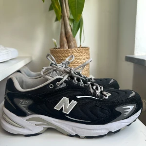 New Balance 725 svarta sneakers - Svarta och grå New Balance 725 sneakers med vita detaljer och klassisk N-logga på sidan. Skorna har snörning, rund tå och platt sula. Ovandelen är i mesh och mocka för en sportig vibe. Perfekta för dig som gillar retrostil och streetwear.