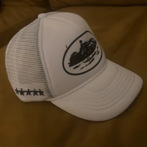 Vit Corteiz truckerkeps med mesh - Vit truckerkeps från Corteiz med svart tryck framtill och broderade stjärnor på sidan. Kepsen har meshpaneler bak för extra ventilation och justerbar snapback. Svart Corteiz-logga broderad bak och detaljerad insida med logoband.