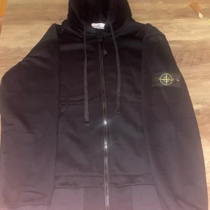  Stone Island zip - Svart hoodie från Stone Island med dragkedja framtill och klassisk logotyp-patch på vänster ärm. Tröjan har huva med snörning och två stora fickor framtill. Tillverkad i mjuk bomull, perfekt för en avslappnad och stilren look.