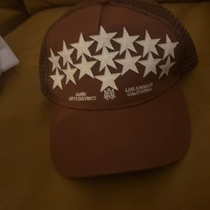 Brun keps från Amiri med stjärnor - Snygg brun keps från Amiri med vit stjärndesign framtill och texten 'AMIRI ARTS DISTRICT LOS ANGELES CALIFORNIA'. Kepsen har mesh på sidorna och justerbar snapback baktill. Perfekt för dig som vill sticka ut med en streetstyle-accessoar.