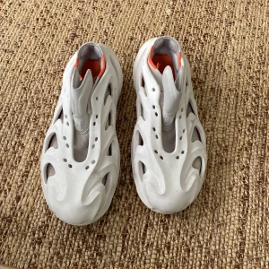 Adidas Adilette 22 vita sneakers - Unika Adidas Adilette 22 sneakers i vitt med futuristisk design och skulpterad sula. Skorna har många utskurna detaljer och orange insida som ger en cool kontrast. Tillverkade i syntetmaterial och passar perfekt för dig som gillar statement pieces. Skosnöre medföljer. Pris kan diskuteras