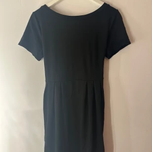 Svart klänning med spetskant - En svart klänning med rund hals och korta ärmar. Kjolen har lätt rynk och nederkanten är dekorerad med snygg spetsdetalj. Klänningen är i mjukt material och har en enkel, stilren look med en feminin touch från spetsen. Storlek M