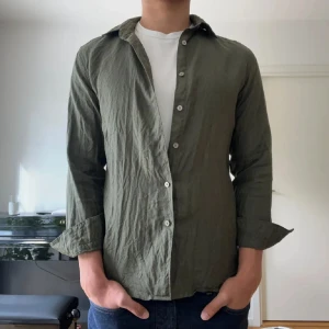 Linneskjorta från Uniqlo  - Snygg olivgrön skjorta i linne med klassisk krage och vita knappar. Skjortan har lång ärm och en avslappnad passform, perfekt att bära öppen över en t-shirt. Materialet är lätt och andas, vilket gör den skön under varmare dagar.