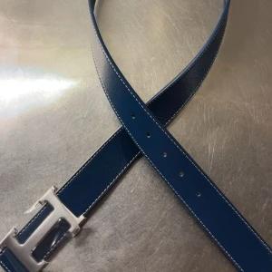 Blått  skärp med silver H-spänne - Snyggt blått skärp  med klassiskt H-spänne i silverfärgad metall. Bältet är gjort i präglat skinn och har vita kontrastsömmar längs kanterna. Perfekt accessoar för att lyfta din outfit med en lyxig touch.