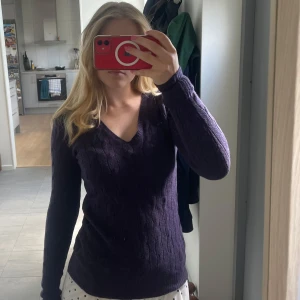 Mörklila stickad tröja Ralph Lauren - Mörklila stickad tröja från Ralph Lauren med v-ringning och kabelstickat mönster. Klassisk grön logga broderad på bröstet. Långärmad och figurnära passform, perfekt för lager-på-lager under höst och vinter.