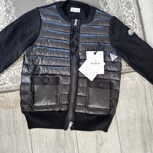 Svart  cardigan  från Moncler - HELT NY ‼️‼️Snygg svart pufferjacka från Moncler med quiltad front och två stora fickor med knappstängning. Jackan har långärmade stickade ärmar och ribbade muddar. Dragkedja framtill och klassisk Moncler-logga på ärmen. Perfekt för dig som vill ha en stilren och sportig look. 