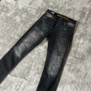 Svarta slim jeans Jack & Jones - Snygga svarta slim fit jeans från Jack & Jones, modell Slim Glenn. Jeansen har slitna detaljer och en tvättad look med fem fickor och bälteshällor. Perfekta för dig som gillar en smal passform och streetinspirerad stil.