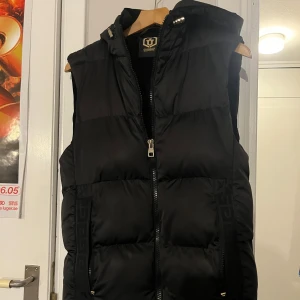 Svart pufferväst från Glorious Gangsta - Säljer en svart dunväst/pufferväst från Glorious Gangsta med huva och dragkedja framtill. Västen är quiltad och har två fickor med diskreta detaljer längs sidorna. funkar till många olika stilar.