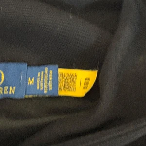 Svart hoodie från Polo Ralph Lauren - Svart hoodie från Polo Ralph Lauren med klassisk broderad logga på bröstet. Tröjan har huva, magficka och ribbade muddar. Tillverkad i mjuk bomull, perfekt för chill dagar eller när du vill ha en clean och stilren look. Storlek M i barn fast passar vuxen i storlek XS