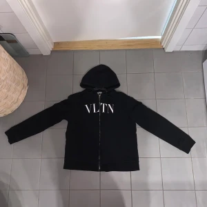 Svart hoodie från Valentino med VLTN - Svart hoodie från Valentino med VLTN-tryck i vitt på bröstet. Klassisk modell med huva, dragkedja och långa ärmar. Tillverkad i mjuk bomull, perfekt för en clean och stilren look. Snygg detalj med broderad logga på insidan.