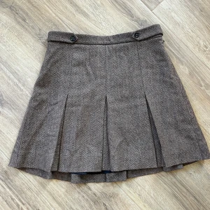 Brun mönstrad kjol från & Other Stories - Snygg brun kortkjol från & Other Stories med fiskbensmönster och veck framtill. Kjolen har två dekorativa knappar i midjan och är fodrad för extra komfort. Perfekt för dig som gillar klassisk men trendig stil.