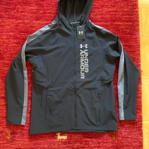 Svart vindjacka från Under Armour - Snygg svart vindjacka från Under Armour med gråa detaljer längs ärmarna och stor logga framtill. Jackan har huva, hel dragkedja och två fickor med dragkedja. Perfekt för träning eller när du vill ha en sportig look. Passar för storlek S också 