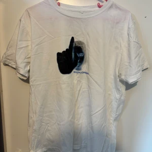 Vit Stella Explorer t-shirt med tryck - Vit t-shirt från Stella Explorer med ett unikt svart tryck av ett ansikte och en hand på bröstet samt texten 'EXPLORER' i blått. Klassisk rund hals och korta ärmar. Skön och mjuk känsla, perfekt för en chill look.