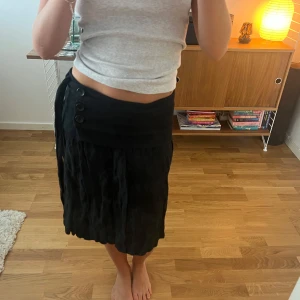 Svart omlottkjol med knappar - Svart omlottkjol med bred midja, tre dekorativa knappar och knytband i sidan. Kjolen har lätt rynkad struktur och slutar strax ovanför knäna. Köpt second hand i Paris 