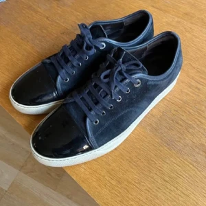 Marinblå Lanvins  - Snygga sneakers i mörkblå mocka med svart, glansig lackad tå och vita sulor. Skorna har rund tå, snörning med svarta skosnören och metallöljetter. Perfekt för dig som gillar en stilren men ändå unik look. Laaanvins