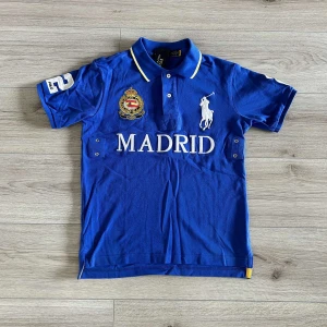 Blå Polo Ralph Lauren Madrid t-shirt - Blå t-shirt från Polo Ralph Lauren med vit text 'MADRID' och stor logga på bröstet. Klassisk pikékrage med gul kant, broderad emblem och stor siffra 2 på ryggen och ärmen. Skön passform och tillverkad i bomull. Perfekt för dig som gillar sportig och stilren look.