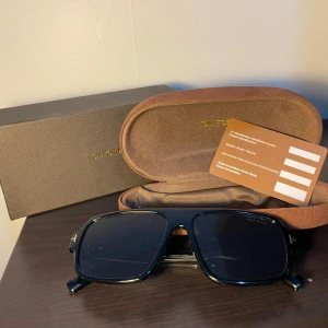  Tom Ford - Snygga svarta solglasögon från Tom Ford med fyrkantig form och breda skalmar. Glasögonen har diskreta gulddetaljer vid gångjärnen och logga på linsen. Kommer med originalfodral och certifikat. Perfekta för dig som vill ha en stilren och lyxig look under sommaren, passar i stort sätt till alla stilar helt nya o använda och allt i bildrn medföljer med glasögonen