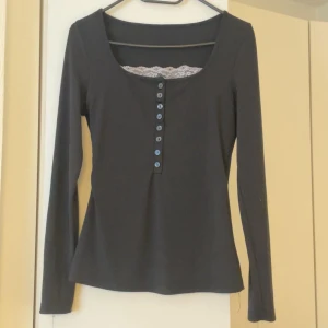 Svart långärmad topp med spetsdetalj - Svart långärmad topp med rundad urringning och dekorativa knappar framtill. Spetsdetalj syns vid halsringningen för en extra touch. Perfekt att styla med jeans eller kjol för en clean look.