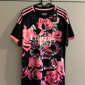 Real Madrid x Adidas rosa/svart fotbollströja - Unik Real Madrid fotbollströja från Adidas med svart bas, rosa rosor och en stor orm på framsidan. Klassiska Adidas-ränder i rosa på axlarna och klubbmärke på bröstet. Tillverkad i lätt och ventilerande polyester.