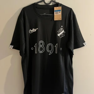 Svart AIK x Nike fotbollströja 1891 - AIK x Nike svart fotbollströja med vit 1891-tryck på bröstet, AIK-emblem och Nike-logga. Kortärmad modell i lätt och ventilerande Dri-FIT-material. Unika rutiga detaljer på ärmsluten och texten 'SMOKINGLIRARE' i nacken. Perfekt för dig som vill representera AIK med stil.