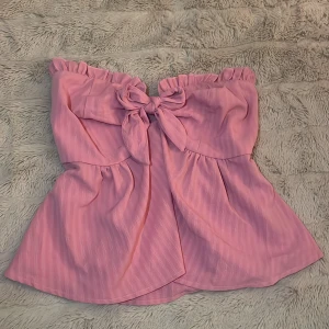 Rosa axelbandslös tubtopp med rosett - Supersöt rosa tubtopp med volangkant upptill och stor rosett framtill. Toppen är axelbandslös och har en lätt utsvängd passform. Perfekt för dig som vill sticka ut med en feminin och trendig look.